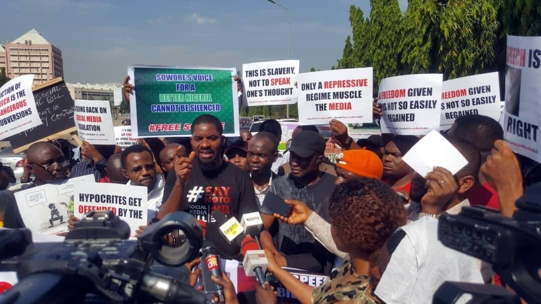 Deji Adeyanju speaking to journalists at a protest, surrounded by placards against the proposed social media bill